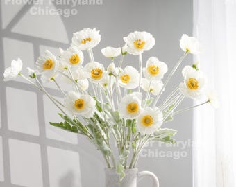 May include: A bouquet of white poppy flowers with yellow centers and green leaves in a white vase. The image includes the text "Flower Fairyland Chicago". The flowers are arranged in a vase with a handle.