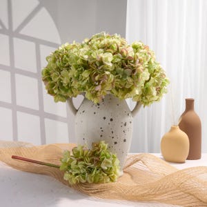 Puede incluir: Arreglo decorativo con flores de hortensia verdes y rosas en un jarr&oacute;n moteado blanco roto con asas. Un tallo de hortensia m&aacute;s peque&ntilde;o y dos jarrones minimalistas crean una est&eacute;tica natural.