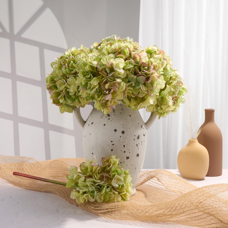 Puede incluir: Arreglo decorativo con flores de hortensia verdes y rosas en un jarr&oacute;n moteado blanco roto con asas. Un tallo de hortensia m&aacute;s peque&ntilde;o y dos jarrones minimalistas crean una est&eacute;tica natural.
