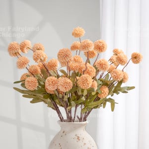 May include: A display of artificial peach-coloured pom-pom flowers with green leaves in a cream-coloured vase. The flowers are spherical and clustered on brown stems. The vase has a textured surface and a wide opening.