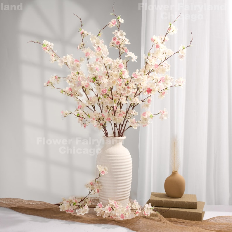 Peut inclure: Un vase en c&eacute;ramique blanc rempli de fausses branches de fleurs de cerisier. Les branches ont de d&eacute;licates fleurs blanches et roses. Un petit vase et des livres sont &eacute;galement visibles. L'arrangement est pos&eacute; sur une surface de ton neutre.