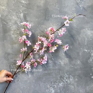 May include: A branch of pink artificial cherry blossoms against a gray textured background. The branch is long and has many small pink flowers.