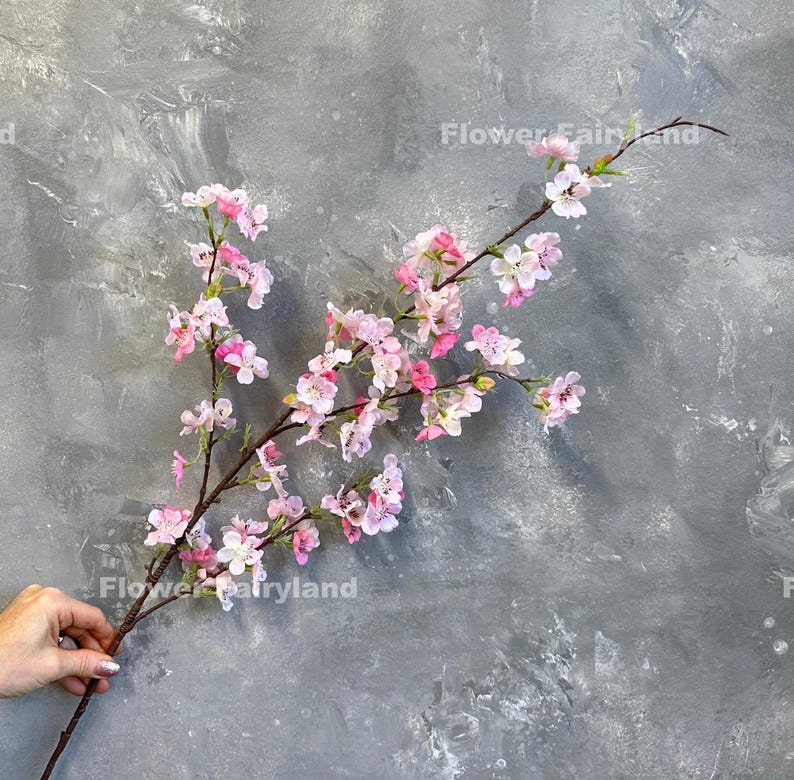 May include: A branch of pink artificial cherry blossoms against a gray textured background. The branch is long and has many small pink flowers.
