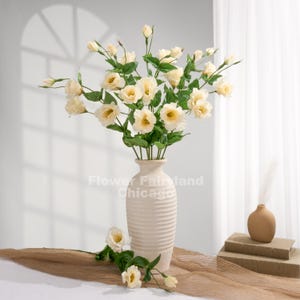 May include: A tall, ribbed, cream-colored ceramic vase holds a large bouquet of pale yellow eustoma flowers with green leaves. The text Flower Fairland Chicago is visible on the vase. A single stem lies at the base of the vase.