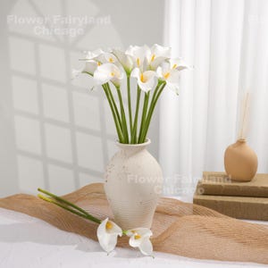 May include: A bouquet of white calla lilies with yellow centers in a cream-colored vase. Two additional lilies rest on a tan fabric. The vase is on a white surface with a neutral background. The text "Flower Fairyland Chicago" is visible.