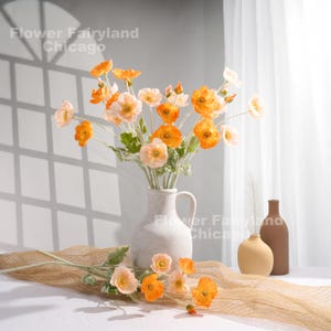 May include: A bouquet of orange and peach artificial poppy flowers in a white ceramic vase. The vase has a textured surface and a spout. The flowers are arranged in a vase on a brown woven surface. The background is a white wall with a window.