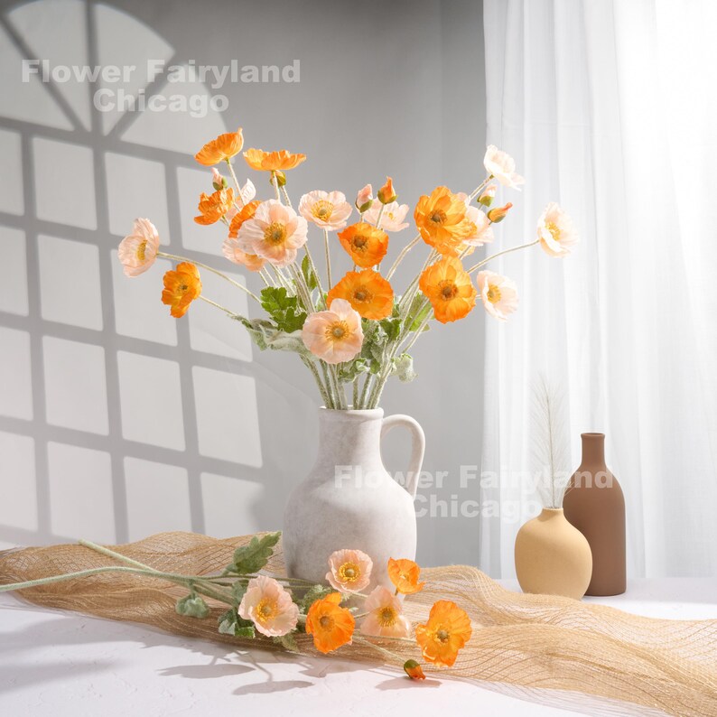 May include: A bouquet of orange and peach artificial poppy flowers in a white ceramic vase. The vase has a textured surface and a spout. The flowers are arranged in a vase on a brown woven surface. The background is a white wall with a window.