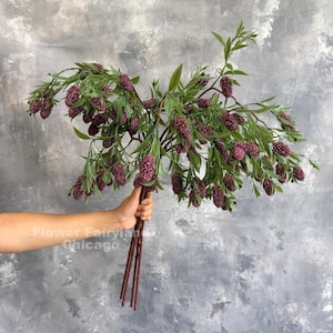 May include: A bundle of artificial branches featuring small, textured, deep purple flower clusters and elongated green leaves. The stems are brown and thin, gathered together at the base. The text Flower Fairyland Chicago is visible.