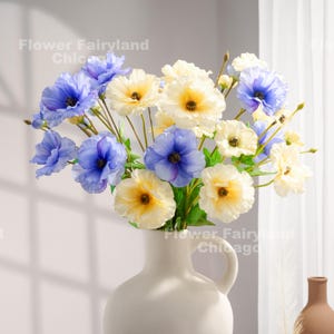 Peut inclure: Un bouquet de fleurs artificielles dans un vase en c&eacute;ramique blanc. Les fleurs sont un m&eacute;lange de bleu et de cr&egrave;me avec un centre jaune. Les fleurs sont dispos&eacute;es dans un vase avec une poign&eacute;e.