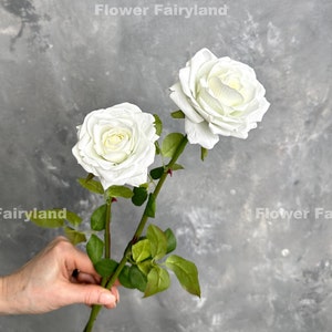 May include: Two white artificial roses with green leaves on a stem. The roses are being held in a hand.