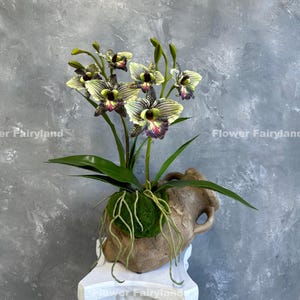 May include: A bouquet of artificial orchids with green and purple flowers in a brown ceramic vase with moss and vines. The vase is sitting on a white pedestal.