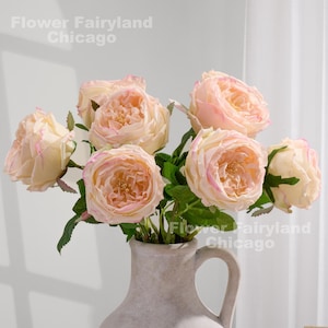 May include: A display of artificial roses in a pale ceramic vase. The roses are a soft peach colour with pink edges, and green leaves. The text "Flower Fairyland Chicago" is visible in the image.