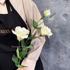May include: A stem of artificial cream-colored eustoma flowers with green leaves and a closed bud. The stem is held against a black apron featuring the text "FAIRYLAND" and "Flower Fairyland Chicago" in light-colored lettering.