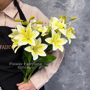 May include: A bouquet of artificial lilies with creamy white petals and vibrant yellow centers, featuring green stems and unopened buds. The black apron worn by the holder displays the text Flower Fairyland Chicago in gold.