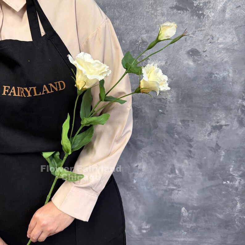 May include: A single stem of artificial lisianthus flowers featuring three creamy white blossoms with ruffled petals and green leaves, held against a black apron with the gold embroidered text FAIRYLAND.