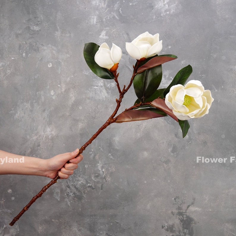 May include: A single stem of artificial magnolia flowers with three white blossoms and green leaves. The stem is brown and the flowers are made of fabric.