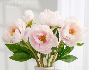 May include: A bouquet of artificial peonies in a ceramic vase. The flowers are a soft pink and white with yellow centers. Green leaves and stems are visible. The vase is a light beige color.
