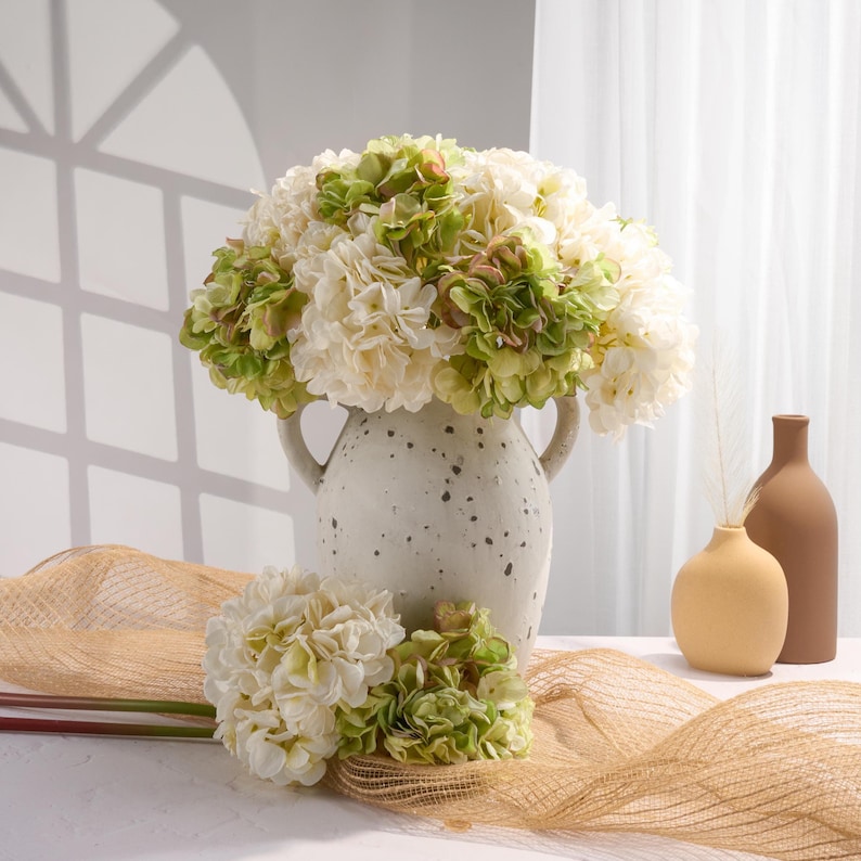 Puede incluir: Un bodeg&oacute;n con un jarr&oacute;n de cer&aacute;mica moteada lleno de flores de hortensia blancas y verdes. Tambi&eacute;n hay dos ramos m&aacute;s peque&ntilde;os y dos jarrones minimalistas. Un camino de mesa de arpillera marr&oacute;n y una superficie blanca completan la escena.