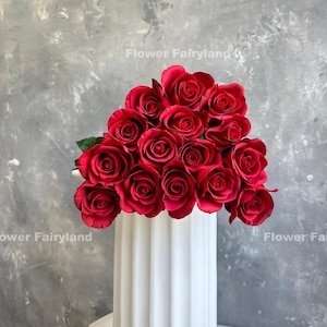 May include: A bouquet of deep red roses arranged in a white vase. The roses are in full bloom and have a velvety texture.
