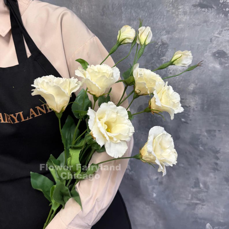 May include: A bouquet of cream-colored lisianthus flowers with ruffled petals, green stems, and leaves. The flowers are held against a black apron with the text Flower Fairyland Chicago visible in gold lettering on the fabric.