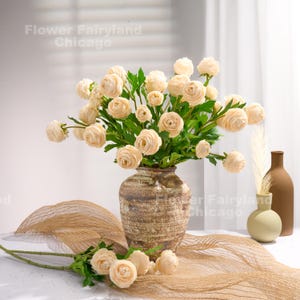 Peut inclure: Un bouquet de fleurs artificielles de renoncules de couleur cr&egrave;me dans un vase en c&eacute;ramique brun. Les fleurs sont dispos&eacute;es dans un vase sur une table recouverte d'un tissu en toile de jute beige.