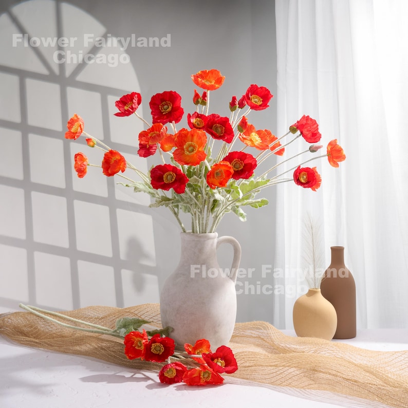 May include: A bouquet of red and orange poppy flowers in a white ceramic vase. The vase has a simple design and a slightly flared opening. The flowers are arranged in a loose, natural style. The background is a light gray wall with a white curtain.