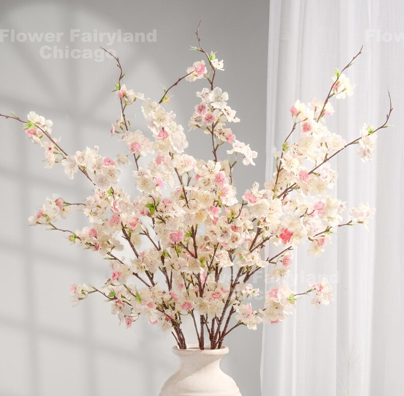 Peut inclure: Un bouquet de branches de fleurs de cerisier artificielles dans un vase blanc. Les branches sont remplies de d&eacute;licates fleurs blanches et roses. Le vase est de conception simple et &eacute;l&eacute;gante. Le texte "Flower Fairyland Chicago" est visible en haut &agrave; gauche.