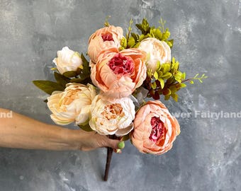 Puede incluir: Un ramo de peonías artificiales en tonos melocotón, crema y rojo. Las flores artificiales tienen múltiples capas de pétalos y están acentuadas con hojas verdes. Los tallos son marrones, sobre fondo gris.