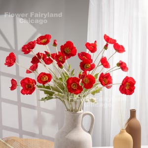 May include: A bouquet of red poppy flowers in a white ceramic vase. The flowers are arranged in a vase with a handle. The vase is sitting on a table with a brown surface.