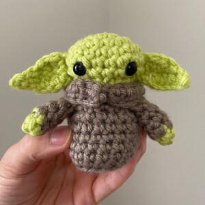 Op de afbeelding: Een gehaakt pluche speelgoed in groen en bruin dat lijkt op een personage uit de Star Wars-franchise. Het speelgoed heeft grote zwarte ogen en een bruin gebreide jas.
