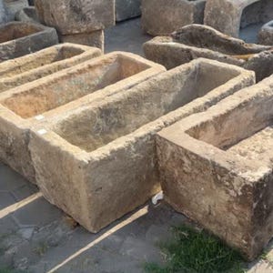 Puede incluir: Una colección de abrevaderos rectangulares de piedra desgastada. Los abrevaderos tienen una superficie rugosa y texturizada en tonos beige y marrón. Las aberturas superiores sugieren que podrían usarse para plantar o con fines decorativos.