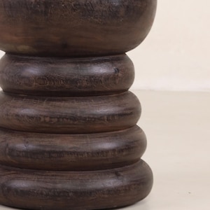 May include: A dark brown wooden pedestal with a rounded top and a series of stacked, rounded rings.