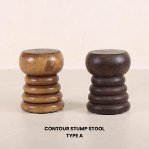 May include: Two wooden contour stump stools, one in a light brown wood and one in a dark brown wood. The stools are round and have a stacked, layered design. The text "CONTOUR STUMP STOOL TYPE A" is below the stools.