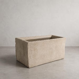 Op de afbeelding: Een rechthoekige plantenbak met een ruw, getextureerd oppervlak in een lichtbeige, steenachtige afwerking. De bak heeft dikke, rechte wanden en een open bovenkant, wat zorgt voor een solide, blokvormig uiterlijk met een neutrale kleur.