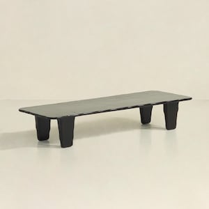 May include: A long, black, rectangular coffee table with a slightly uneven surface and four thick, black legs.