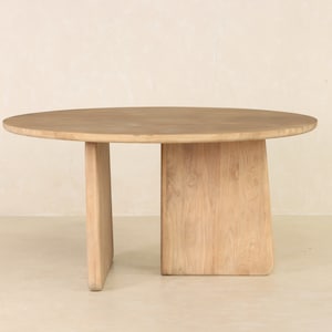 May include: A light-colored, round wooden table with a minimalist design. The table has a circular top supported by two rectangular legs. The legs are connected by a solid, vertical panel. The table is made of natural wood.