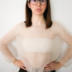 Könnte beinhalten: Eine Frau trägt einen weißen, durchsichtigen Strickpullover mit einem floralen Stirnband und einer schwarzen Brille.