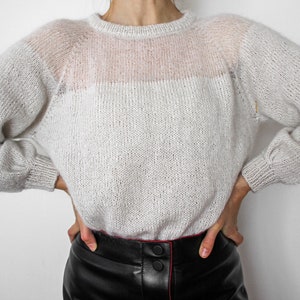 Könnte beinhalten: Ein hellgrauer Strickpullover mit leicht durchsichtiger Textur. Der Pullover hat einen runden Ausschnitt und lange Ärmel.