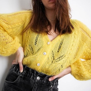 May include: A yellow knitted cardigan with a V-neck and button closure. The cardigan has a delicate openwork pattern and a slightly oversized fit.