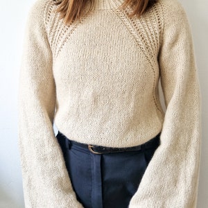 Könnte beinhalten: Ein beiger Strickpullover mit hohem Kragen und Glockenärmeln. Der Pullover hat ein zartes Spitzenmuster an den Schultern.