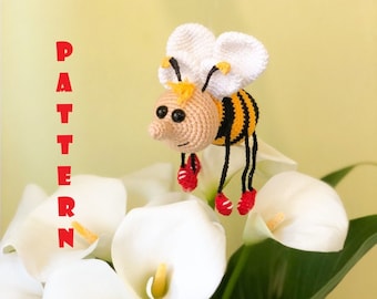 May include: A crocheted bee toy with black and yellow stripes, white wings, and red shoes. The bee is sitting on white calla lilies.