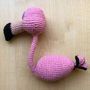 May include: Pink crocheted flamingo toy with black eyes and a black beak. The flamingo is standing on a wooden surface.