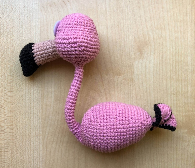 May include: Pink crocheted flamingo toy with black eyes and a black beak. The flamingo is standing on a wooden surface.