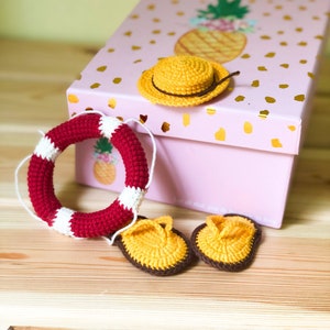 May include: A pink gift box with a pineapple design on the lid. The box is open and contains a crocheted yellow hat, a red and white crocheted life preserver, and two yellow crocheted sandals.