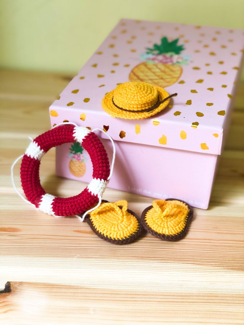 May include: A pink gift box with a pineapple design on the lid. The box is open and contains a crocheted yellow hat, a red and white crocheted life preserver, and two yellow crocheted sandals.