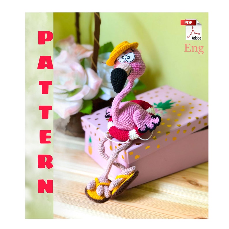 May include: A pink crocheted flamingo wearing a yellow hat and a red and white life preserver. The flamingo is sitting on a pink box with gold pineapple designs. The flamingo has large, black eyes and a black beak. The image includes the text "PATTERN" and "Eng".