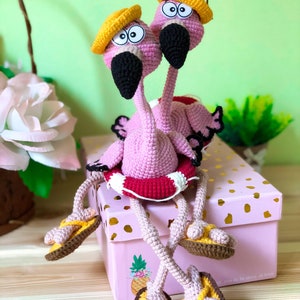 May include: Two pink crocheted flamingo toys wearing yellow hats and brown sandals, sitting on a pink box with gold polka dots and a pineapple design. The flamingoes are holding a red and white life preserver.