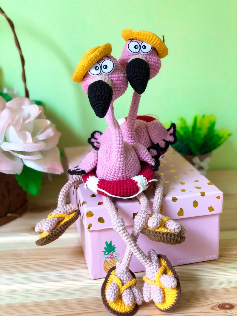 May include: Two pink crocheted flamingo toys wearing yellow hats and brown sandals, sitting on a pink box with gold polka dots and a pineapple design. The flamingoes are holding a red and white life preserver.