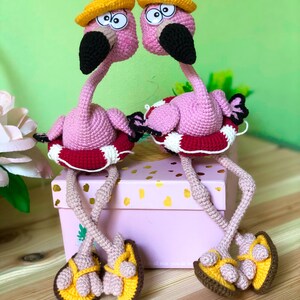 May include: Two pink crocheted flamingo toys wearing yellow hats and red and white life preservers. The flamingos are sitting on a pink box with the text "all that you do be" on the front. The flamingos are wearing yellow and brown crocheted sandals.