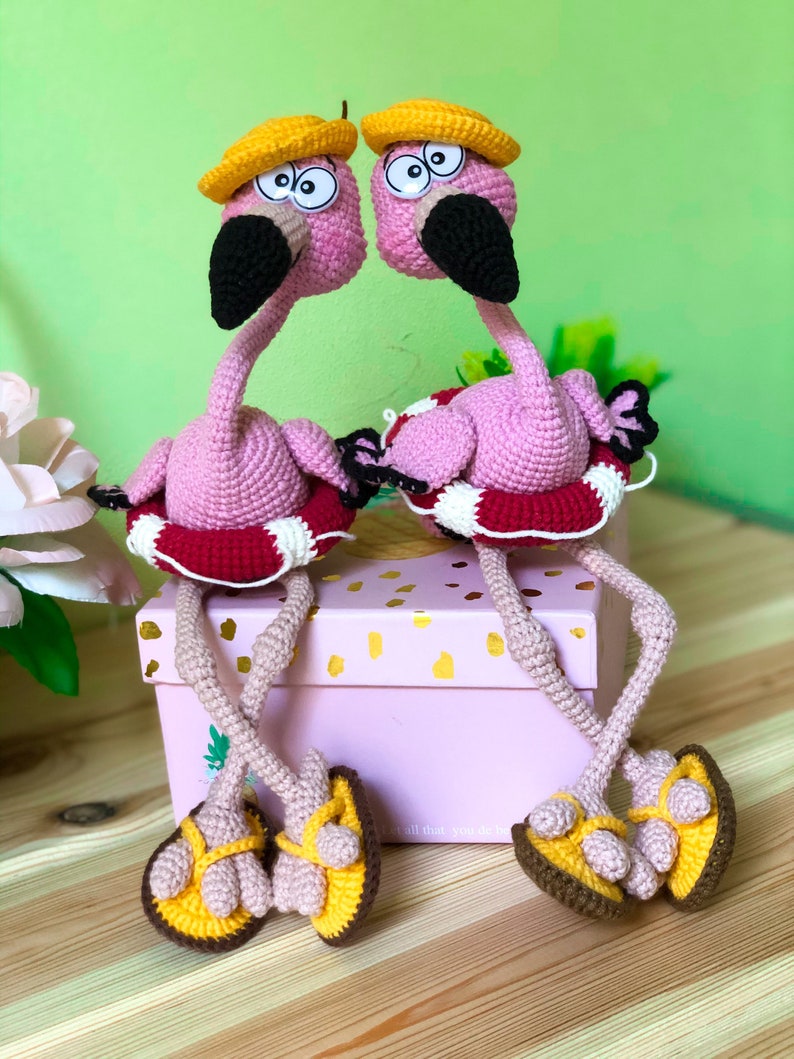 May include: Two pink crocheted flamingo toys wearing yellow hats and red and white life preservers. The flamingos are sitting on a pink box with the text "all that you do be" on the front. The flamingos are wearing yellow and brown crocheted sandals.